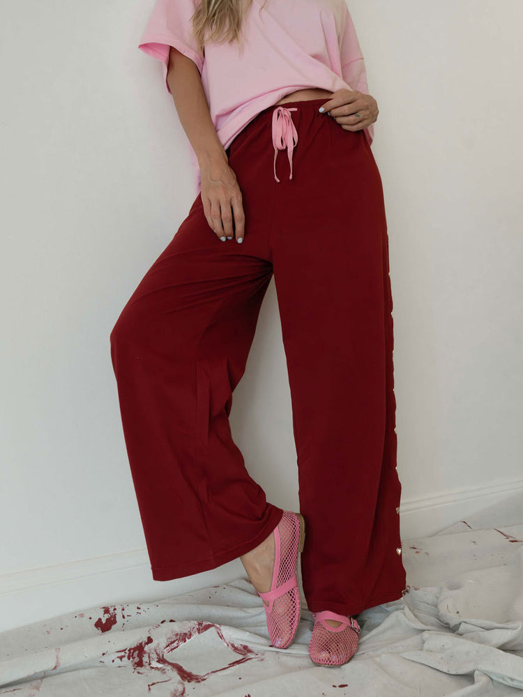 A model wearing loose, wide-leg burgundy pants with a pink drawstring waist, paired with a pink t-shirt and pink mesh flats, standing against a white wall.