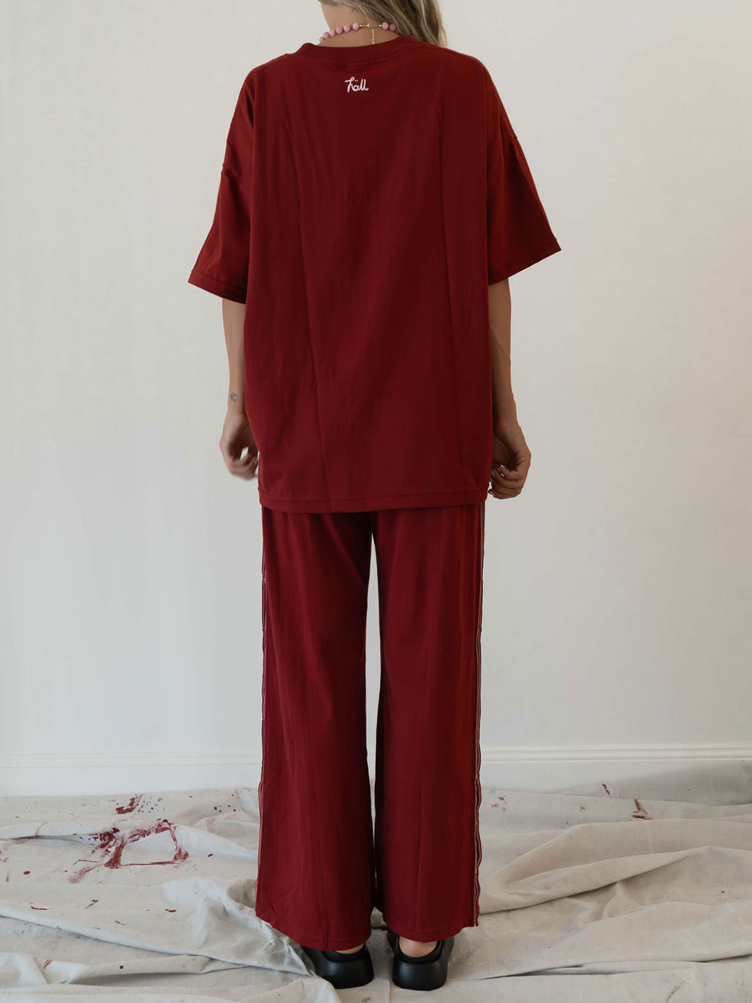 A person shown from the back wearing an oversized deep red short-sleeve top and matching wide-leg pants, standing against a plain off-white wall with a light fabric sheet on the floor.
