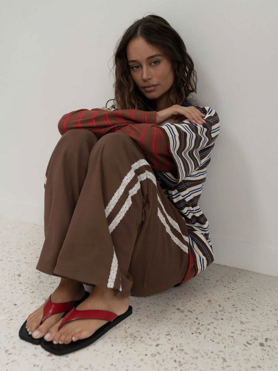 A woman sits on a speckled floor against a white wall, wearing brown wide-leg sweatpants with white lace stripe trim details, a red and brown striped long-sleeve top, and red-and-black flip-flops.