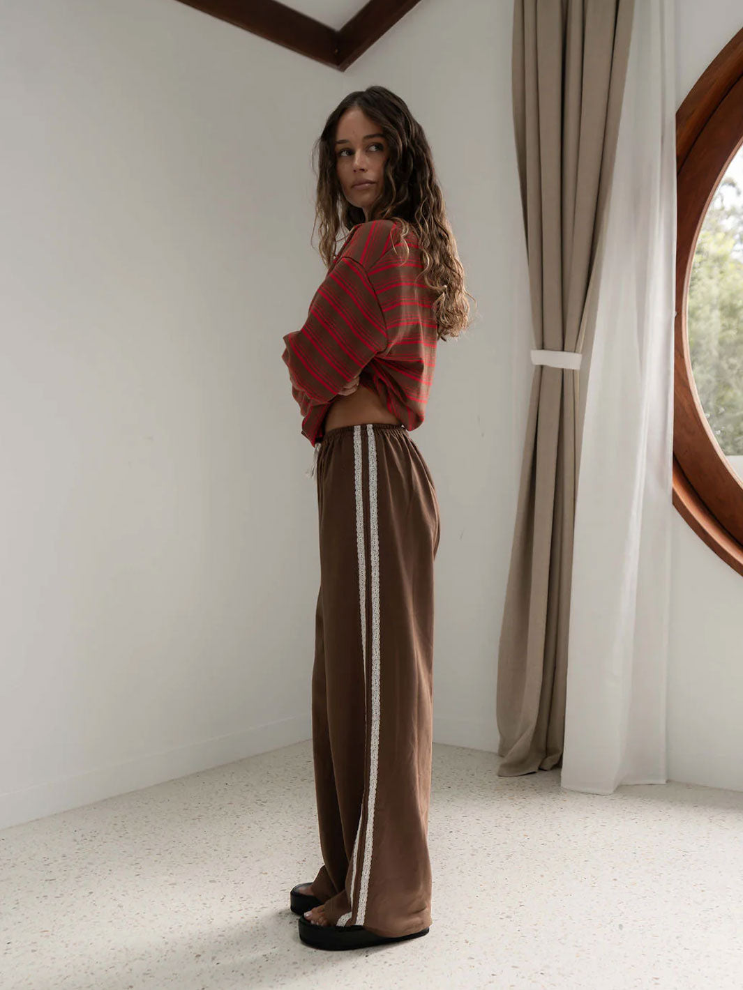 A woman stands in a minimalist room with white walls and a large oval window, wearing a red and brown striped long sleeve t-shirt and brown wide-leg pants with white lace side stripes.