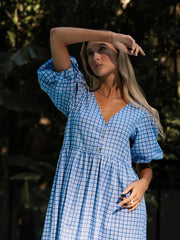 Woman wearing a blue checkered dress standing outdoors with greenery in the background