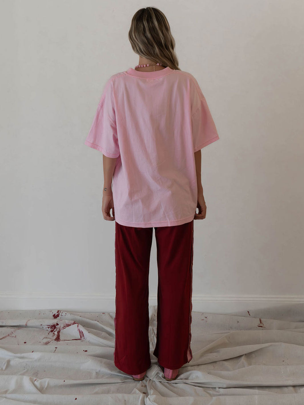 A woman stands with her back to the camera wearing an oversized light pink t-shirt and dark red wide-leg pants, standing on a white sheet with red paint splatters against a plain off-white wall.