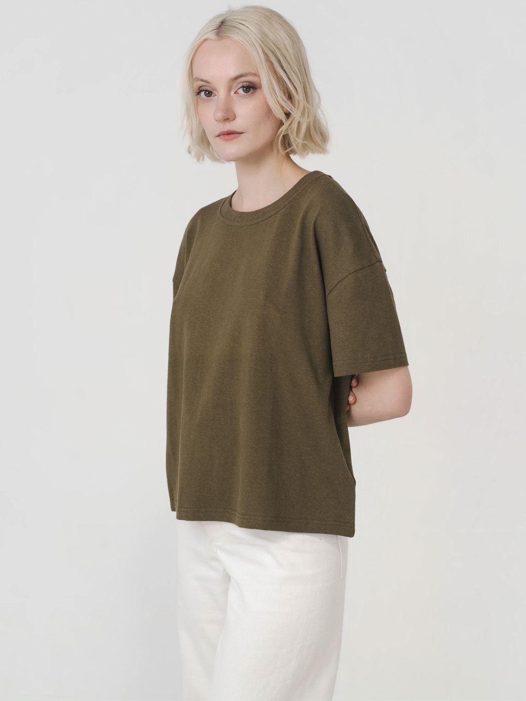A woman with short blonde hair wearing an oversized olive green short-sleeve t-shirt and white pants, standing against a plain light gray studio background.