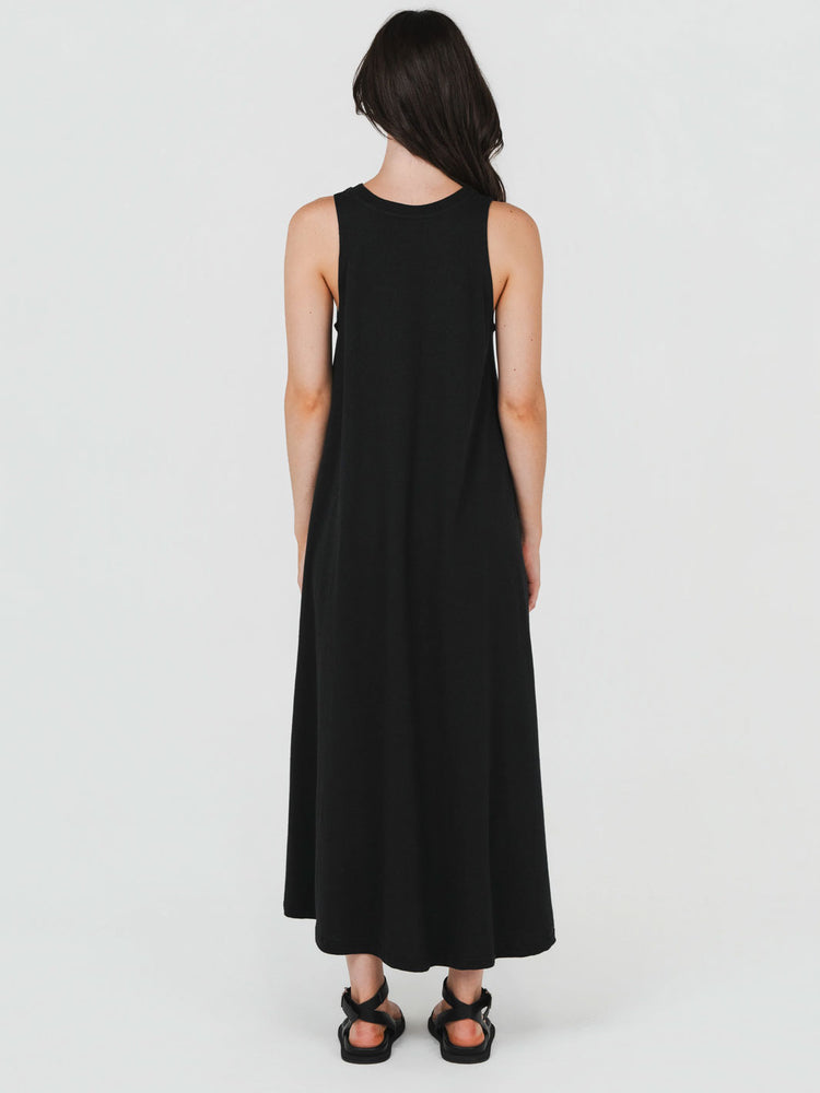 A woman viewed from the back wearing a sleeveless black maxi dress and black strappy sandals against a plain light gray studio background.
