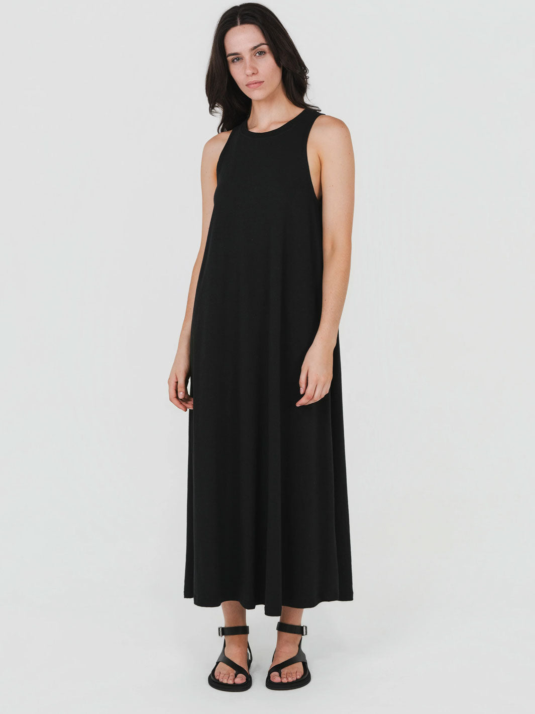 A woman modeling a sleeveless black maxi dress with a relaxed, flowing silhouette and black strappy sandals, set against a plain light gray studio background.
