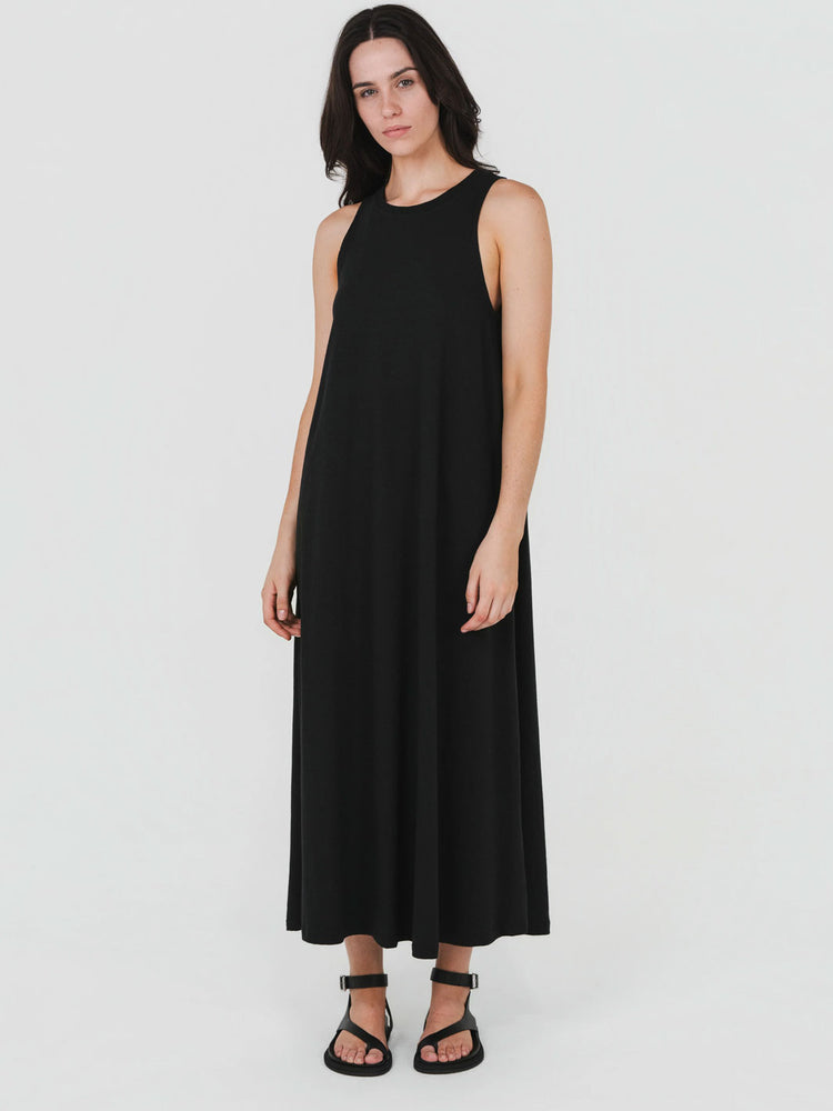 A woman modeling a sleeveless black maxi dress with a relaxed, flowing silhouette and black strappy sandals, set against a plain light gray studio background.