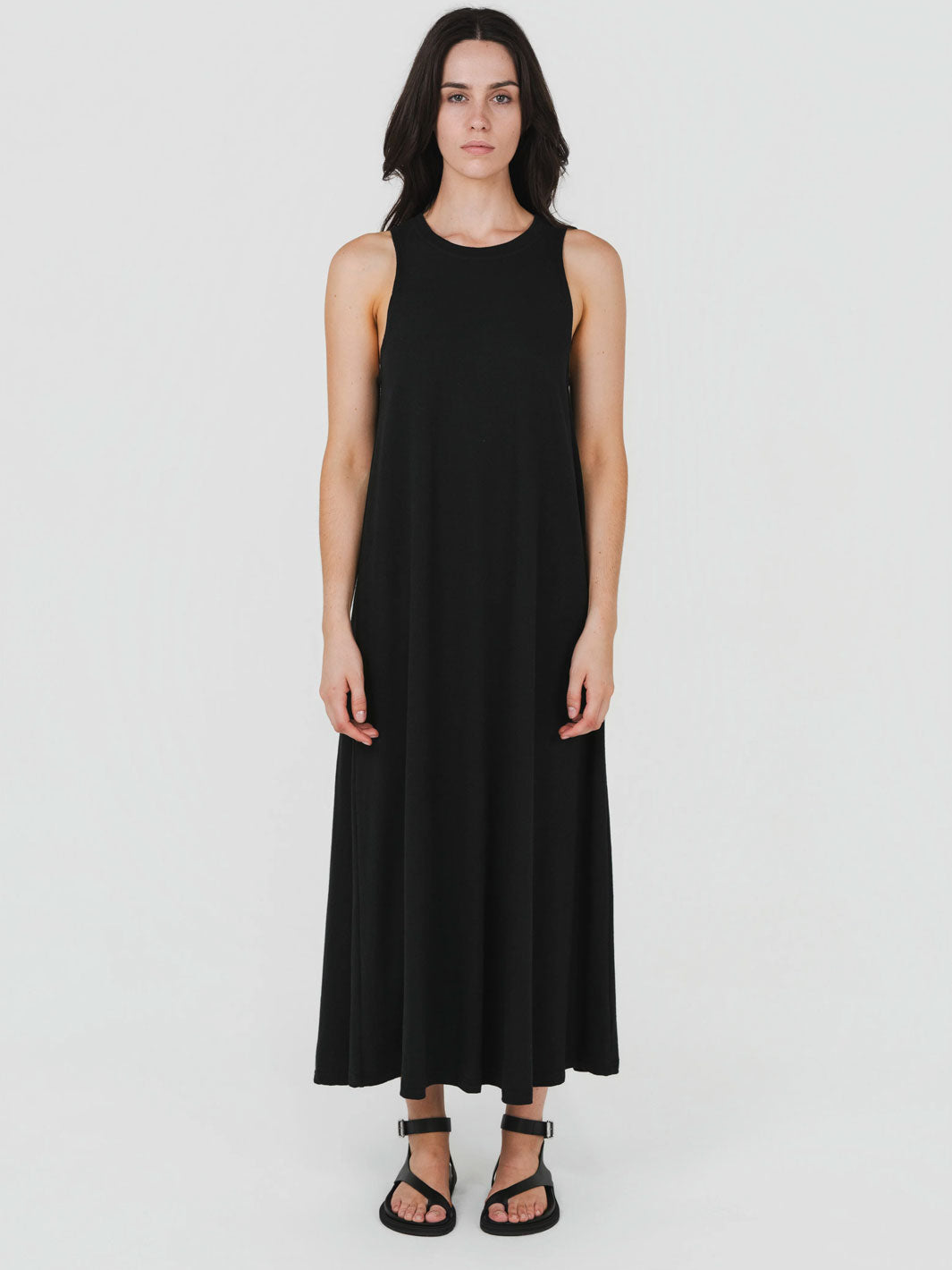 A woman modeling a sleeveless black maxi dress with a relaxed, flowing silhouette, paired with black strappy sandals, against a plain light gray studio background.
