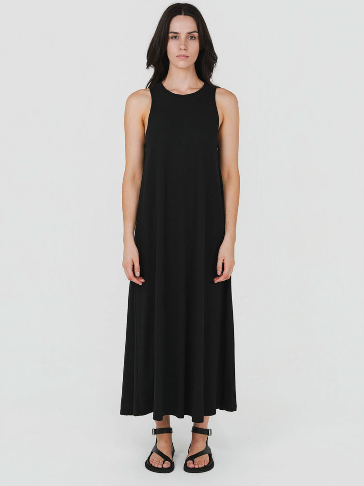 A woman modeling a sleeveless black maxi dress with a relaxed, flowing silhouette, paired with black strappy sandals, against a plain light gray studio background.