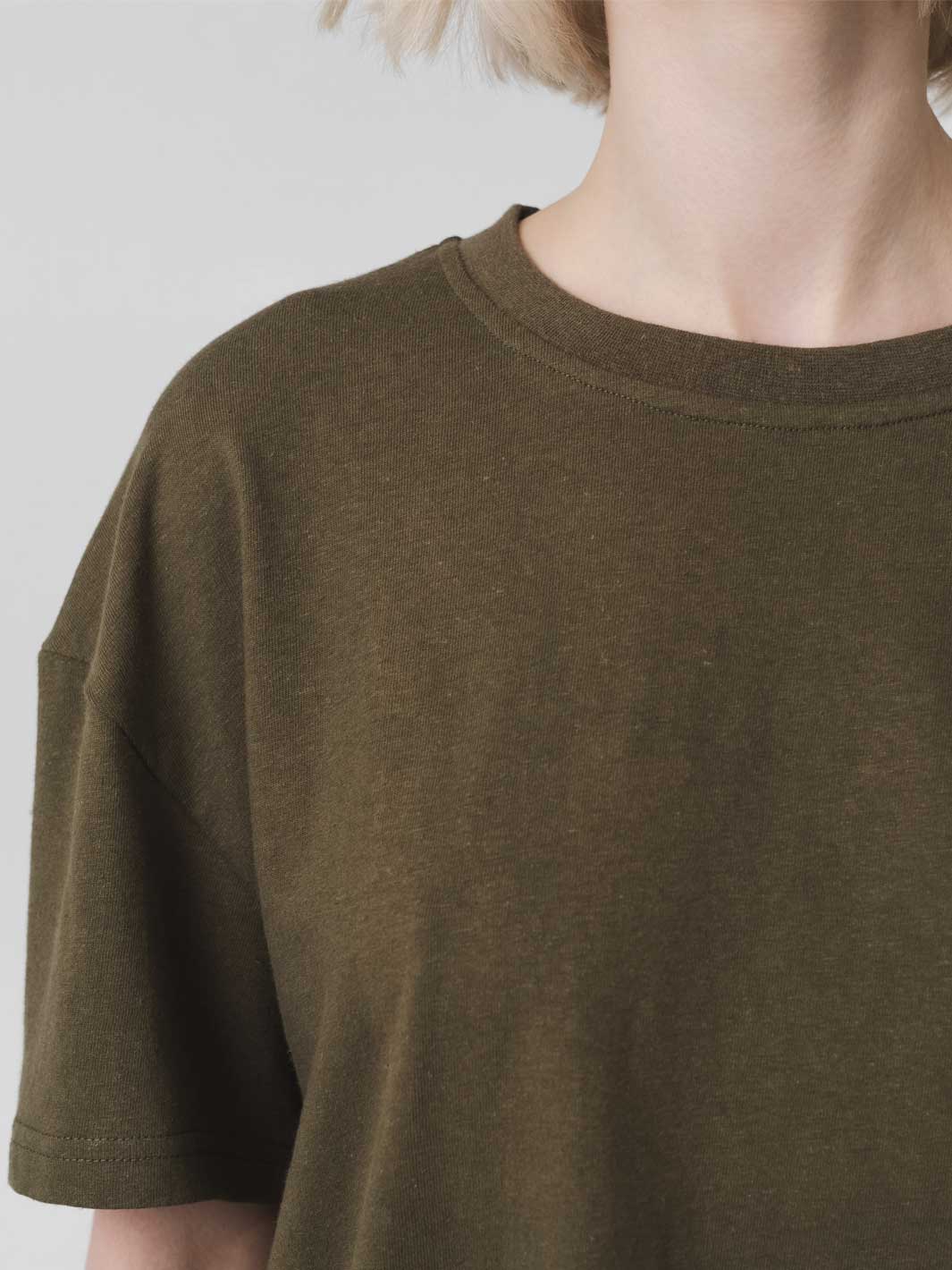 Close-up of a person wearing an oversized short-sleeve crewneck t-shirt in olive green, showing the neck, collar, and shoulder area against a plain light gray background.