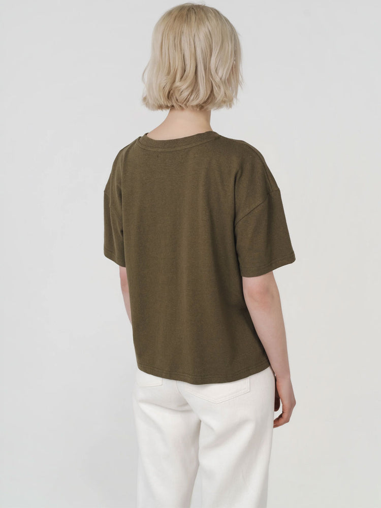 A person shown from the back wearing an oversized olive green short-sleeve t-shirt and white pants, photographed against a plain light gray studio background.
