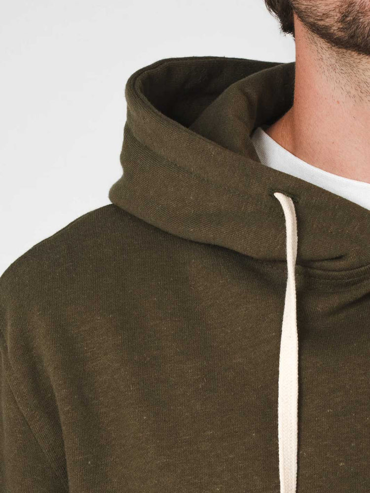 Close-up of a person wearing an olive green hemp and organic cotton hooded sweatshirt with cream-colored drawstrings over a white undershirt, against a plain light gray background.