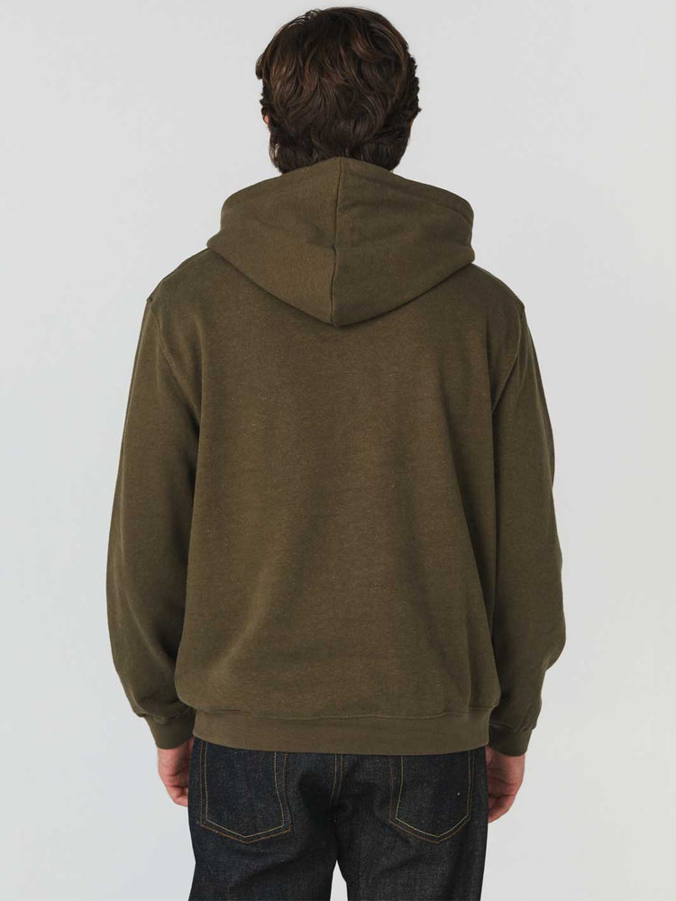 A man viewed from the back wearing an olive green hemp/organic cotton pullover hoodie and dark blue jeans against a plain light gray studio background.
