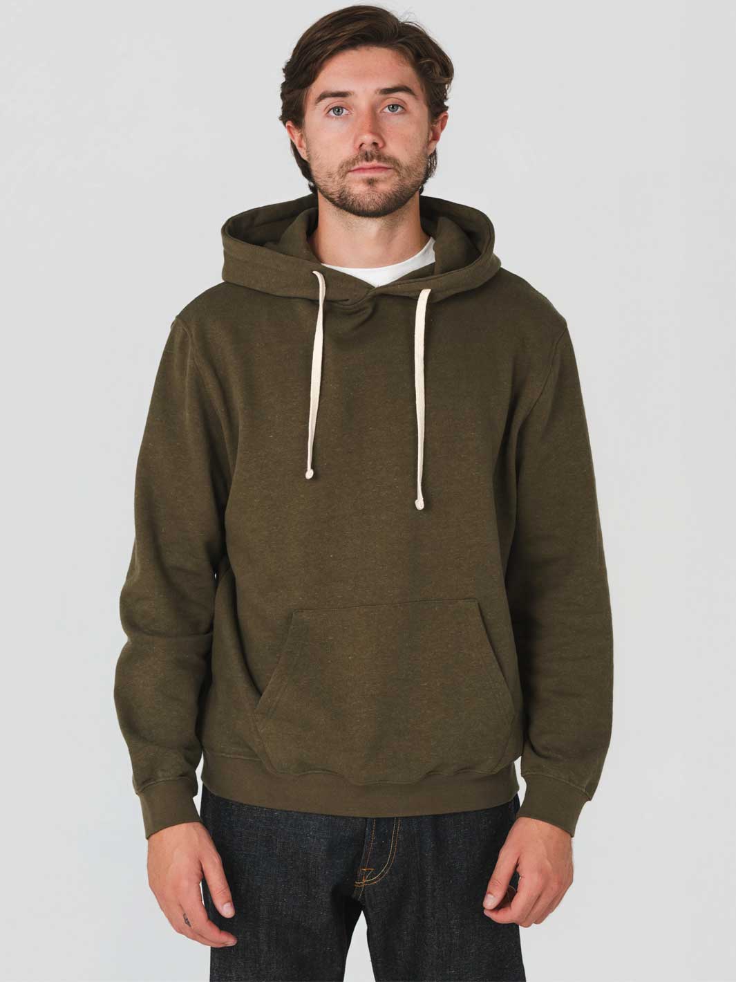 A man wearing an olive green pullover hoodie with cream-colored drawstrings, paired with dark jeans, against a plain light gray studio background.
