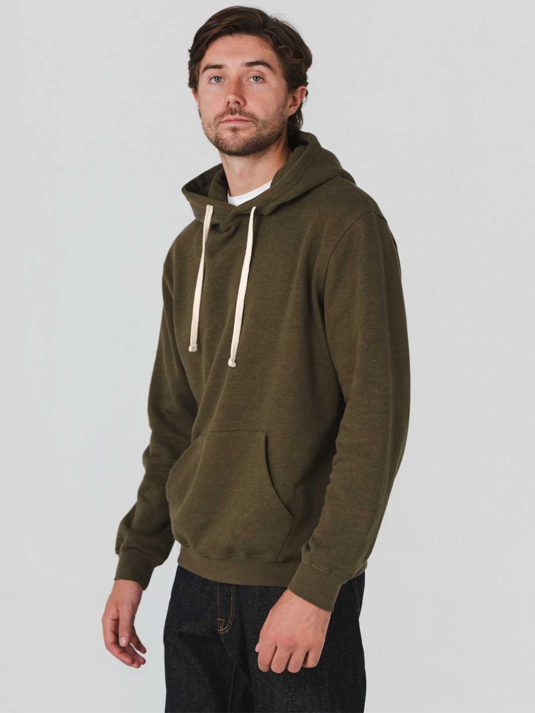 A man wearing an olive green hemp/ organic cotton pullover hoodie with cream-colored drawstrings, paired with dark jeans, photographed against a solid light gray studio background.