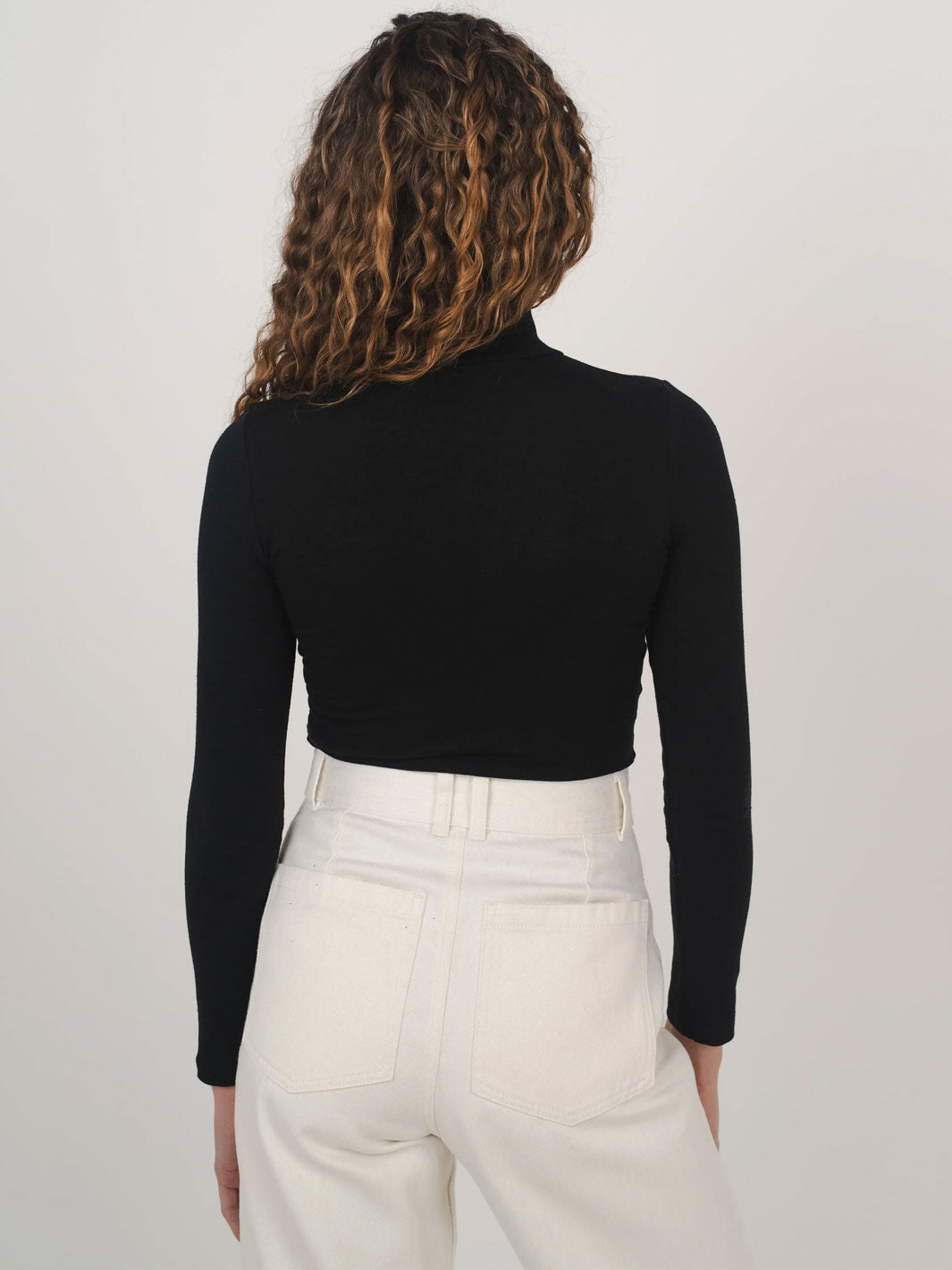 A woman shown from the back wearing a fitted black long-sleeve mock neck skivvy top and high-waisted off-white trousers, in natural materials, against a plain light gray studio background.