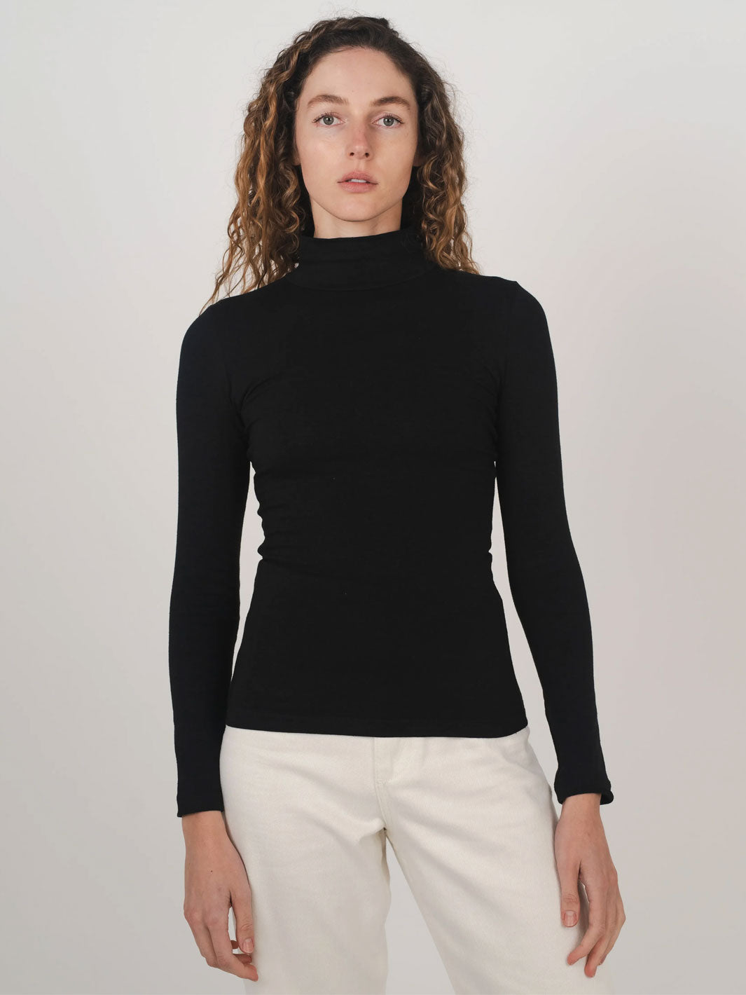 A woman wearing a fitted black long-sleeve mock neck skivvy top and off-white trousers, standing against a plain light gray studio background.