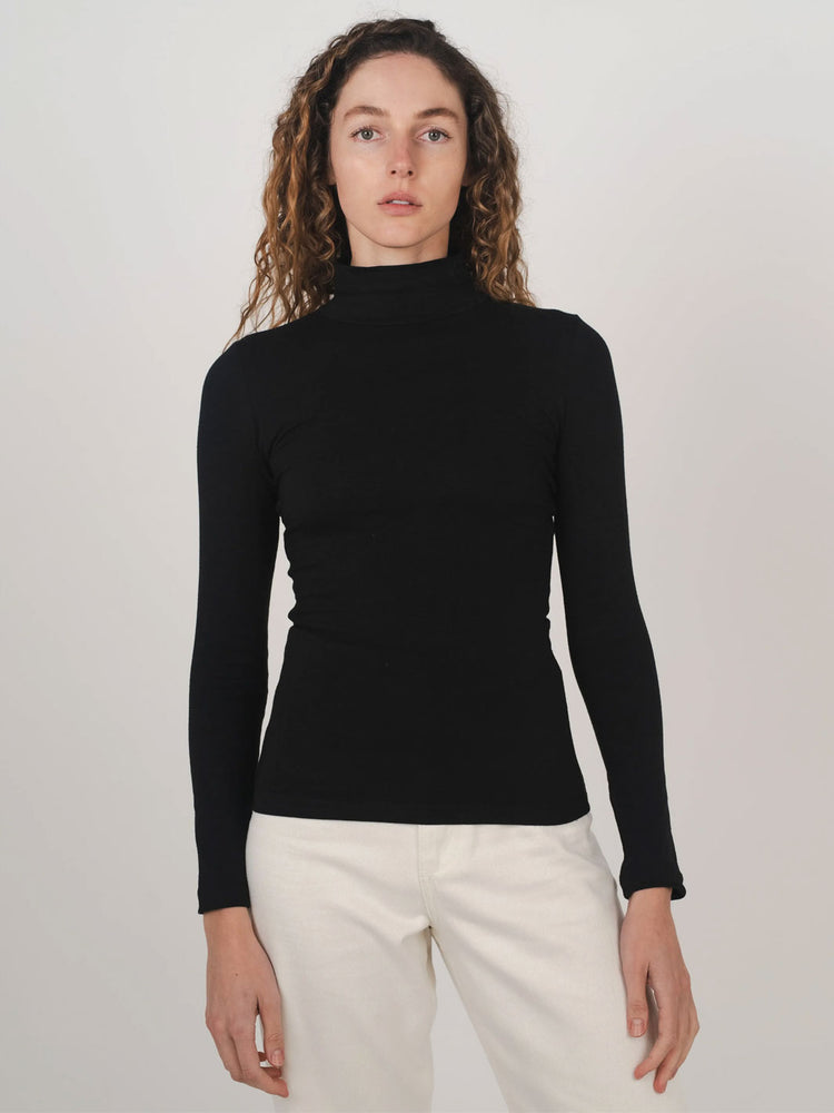 A woman wearing a fitted black long-sleeve mock neck skivvy top and off-white trousers, standing against a plain light gray studio background.