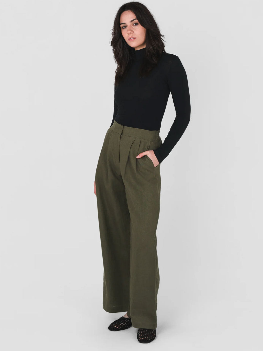 A woman modeling a black long-sleeve mock neck top and olive green wide-leg trousers against a light gray studio background.