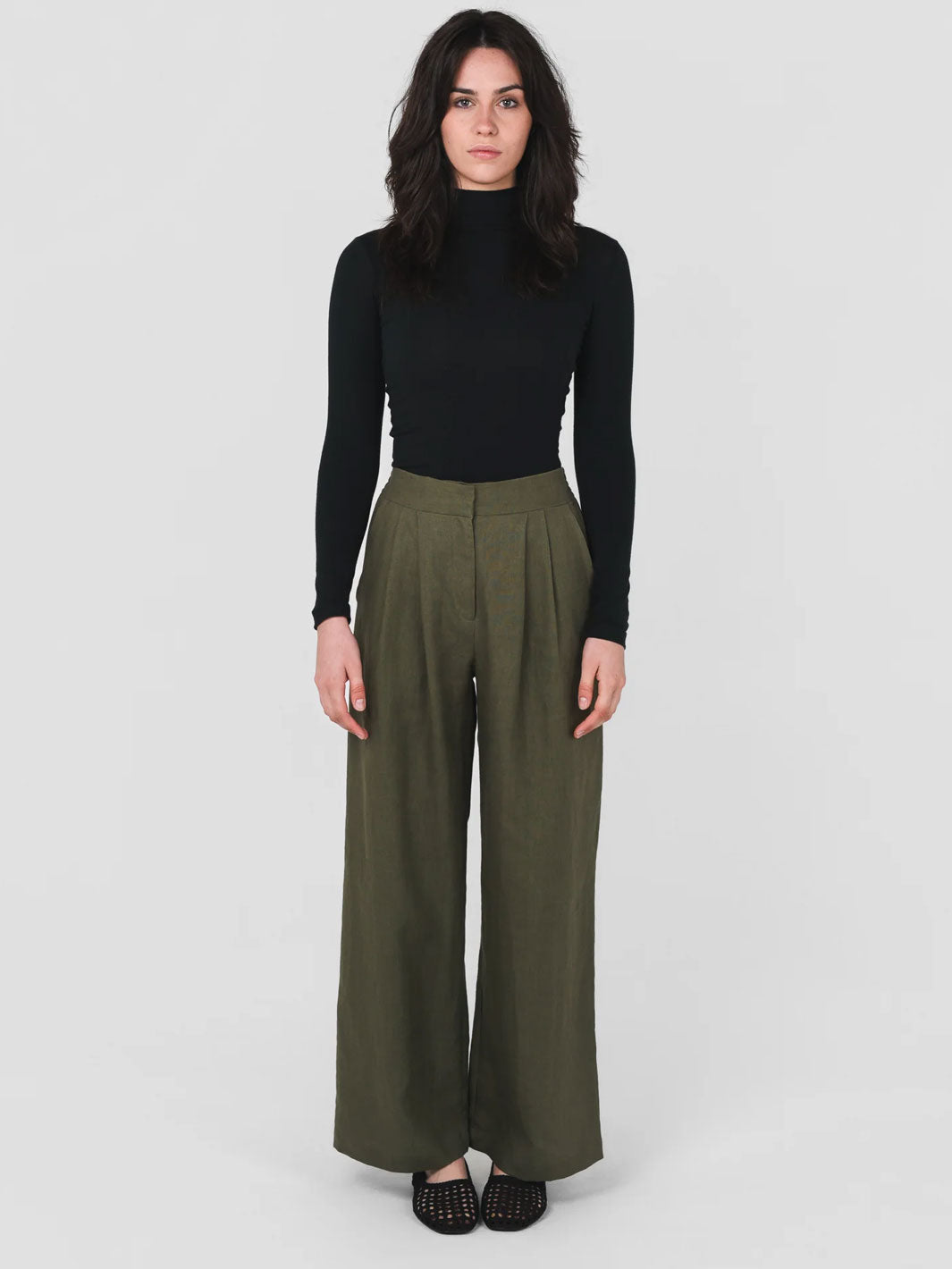 A woman modeling professional workwear attire in natural fibre. A black long-sleeve mock neck top and olive green wide-leg trousers against a light gray studio background.