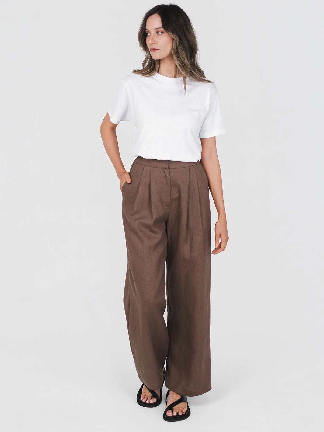 Woman wearing a white t-shirt and brown hemp trousers with pleated front on a white background
