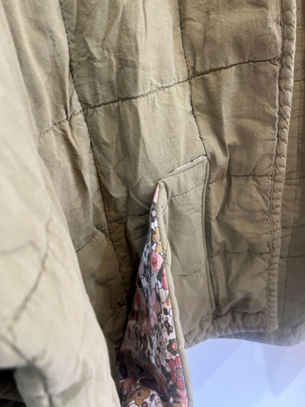 Close-up of a quilted jacket in a warm khaki-beige color, showing the interior lining with a pink, red, and white floral pattern.