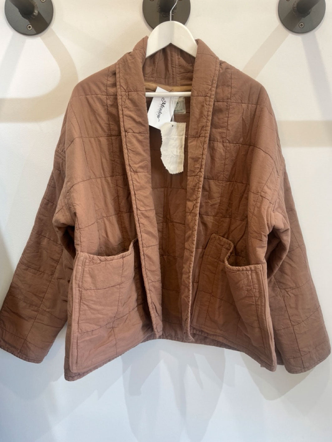 A brown quilted jacket with a shawl collar and large front patch pockets, hanging on a white hanger against a white wall.