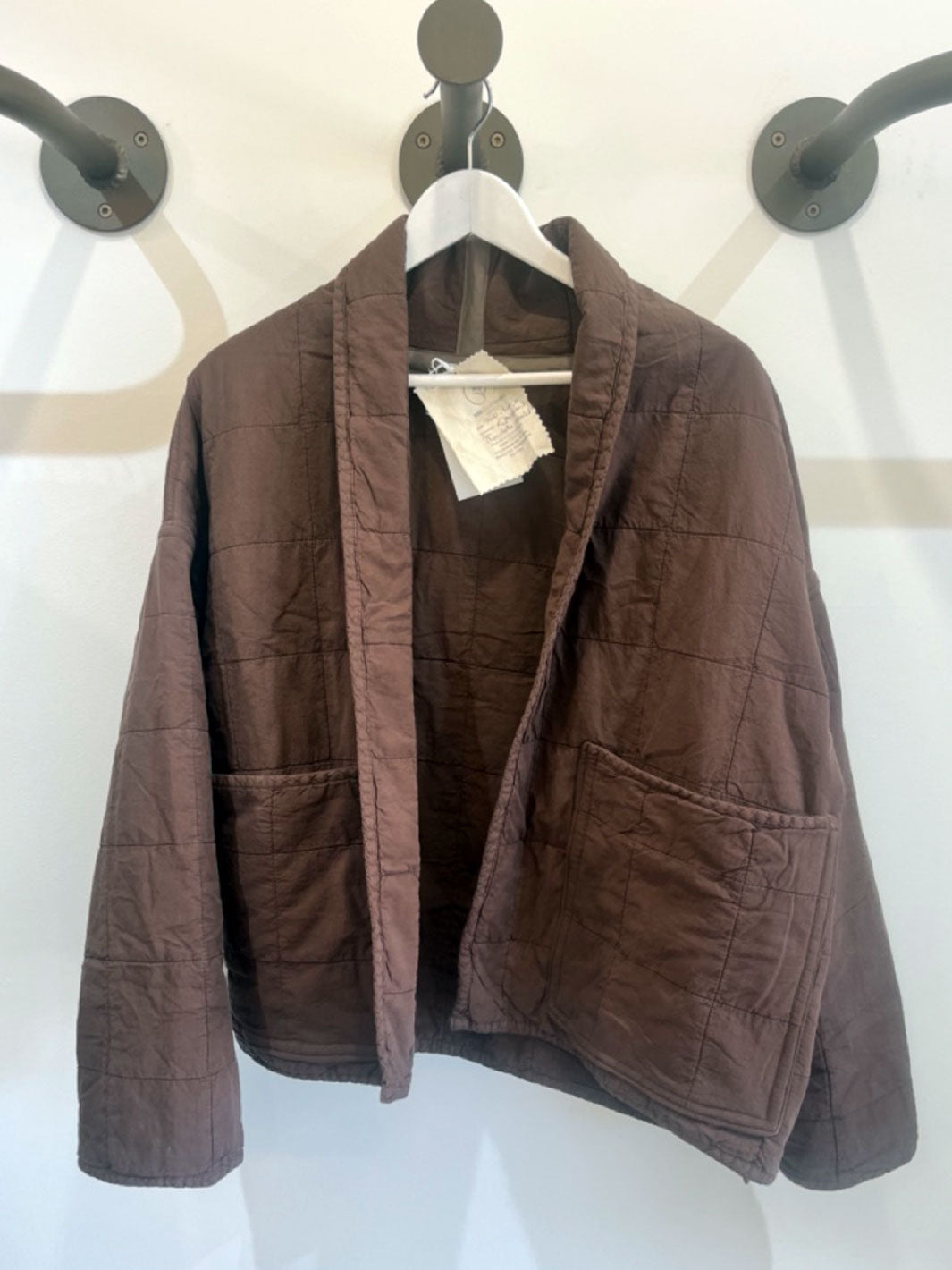 A brown quilted jacket with a shawl collar and front patch pockets, hanging on a white hanger against a white wall.