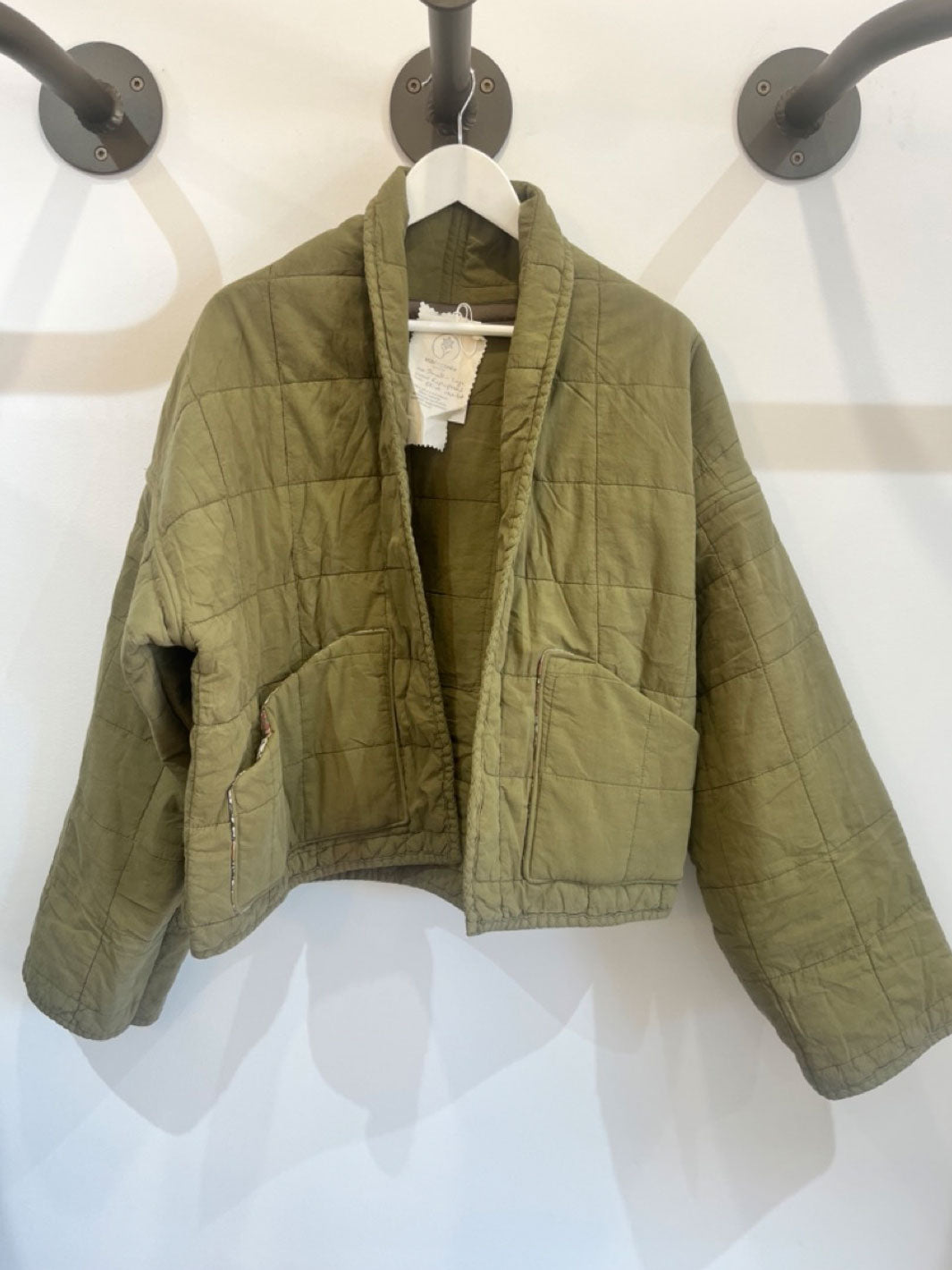 An olive green quilted jacket hanging on a white hanger against a white wall with black wall hooks.