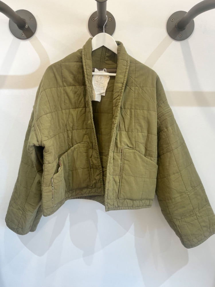 An olive green quilted jacket hanging on a white hanger against a white wall with black wall hooks.