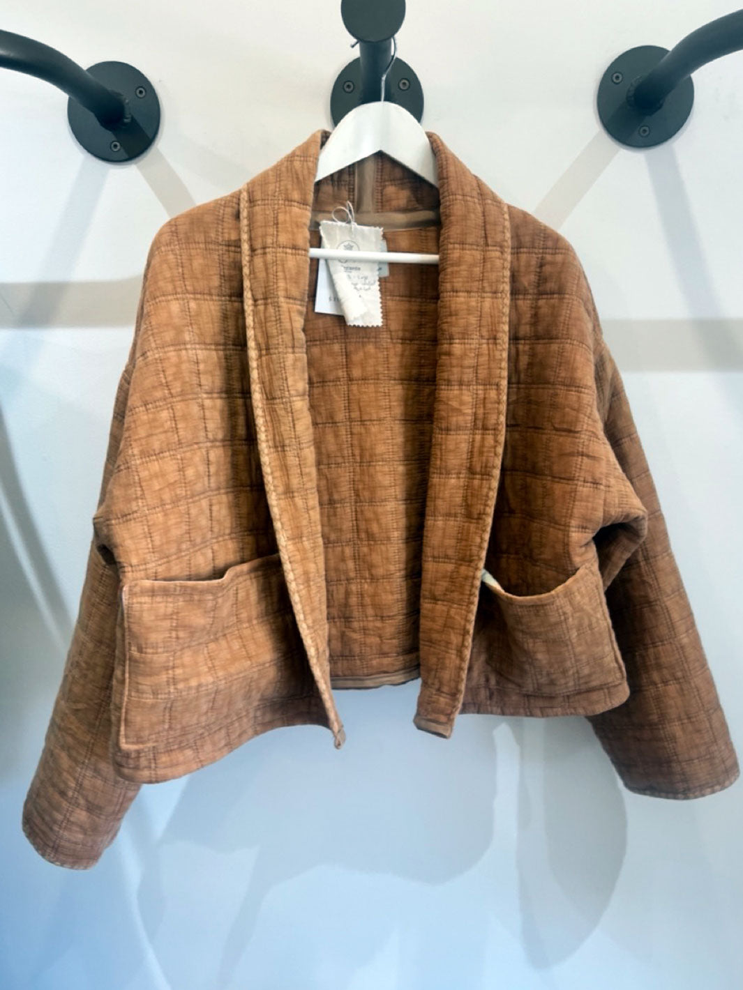 A cropped brown jacket with a shawl collar and front patch pockets, hanging on a white hanger against a white wall.