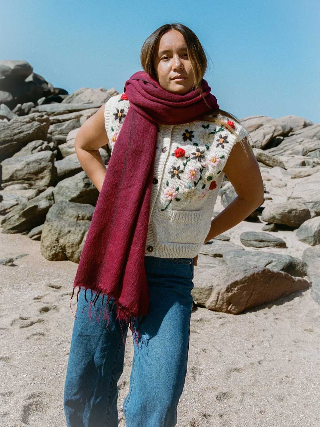 A woman standing outdoors in front of large rocks under a blue sky, wearing a cream knit vest with colorful floral embroidery, blue jeans, and a deep burgundy scarf.