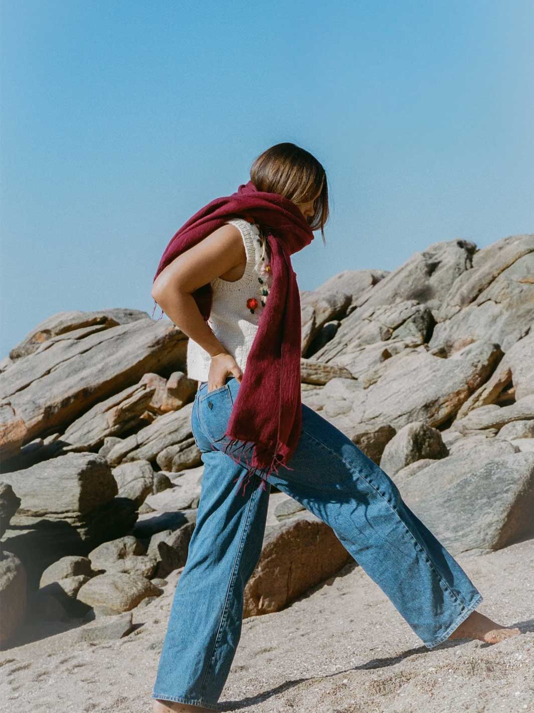 A woman standing on a rocky beach wearing a white sleeveless top, wide-leg blue jeans, and a deep red scarf, with a clear blue sky behind her.