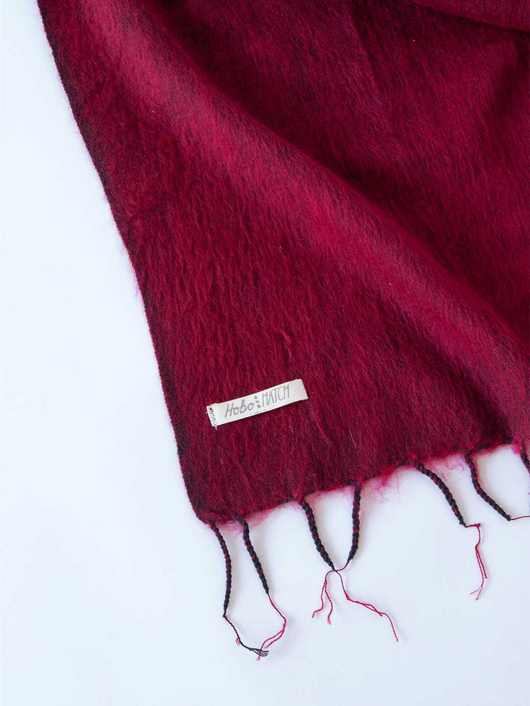A close-up of a deep burgundy fuzzy scarf with black and pink fringe tassels, featuring a small white fabric label that reads 'Mehle & Hille'.