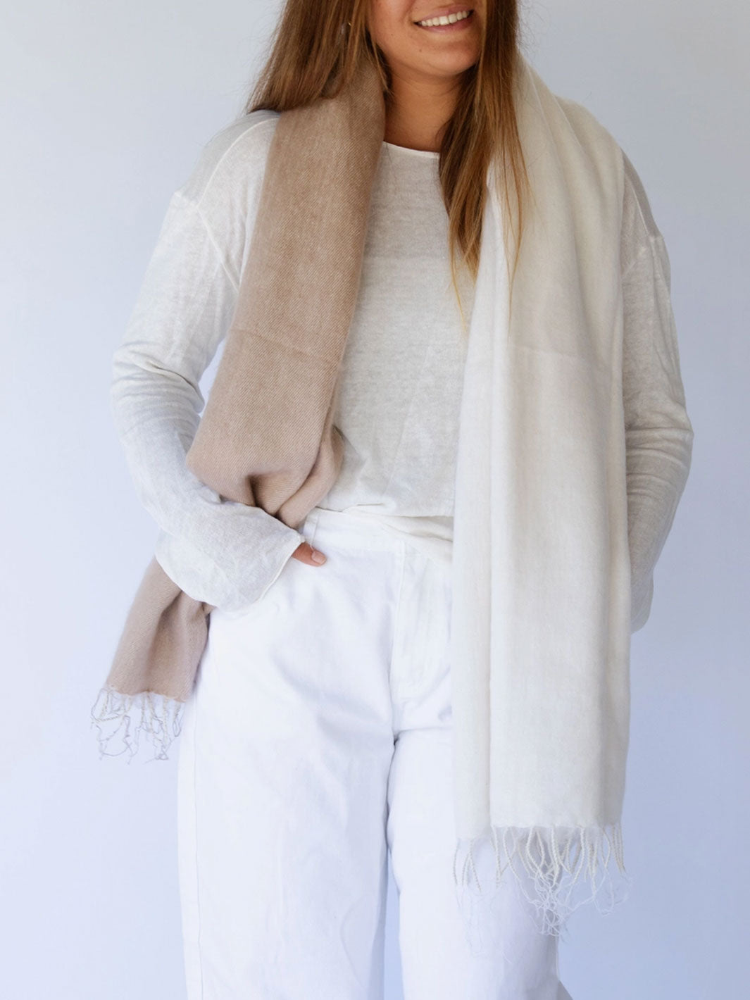 A woman wearing a light beige knit sweater, white pants, and a cream-colored fringed scarf draped over her shoulders, set against a plain light gray background.
