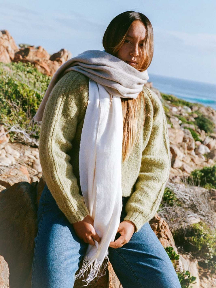 A woman sitting outdoors on a rocky hillside near the ocean, wearing a chunky olive green knit sweater, blue jeans, and a long cream scarf draped around her neck.