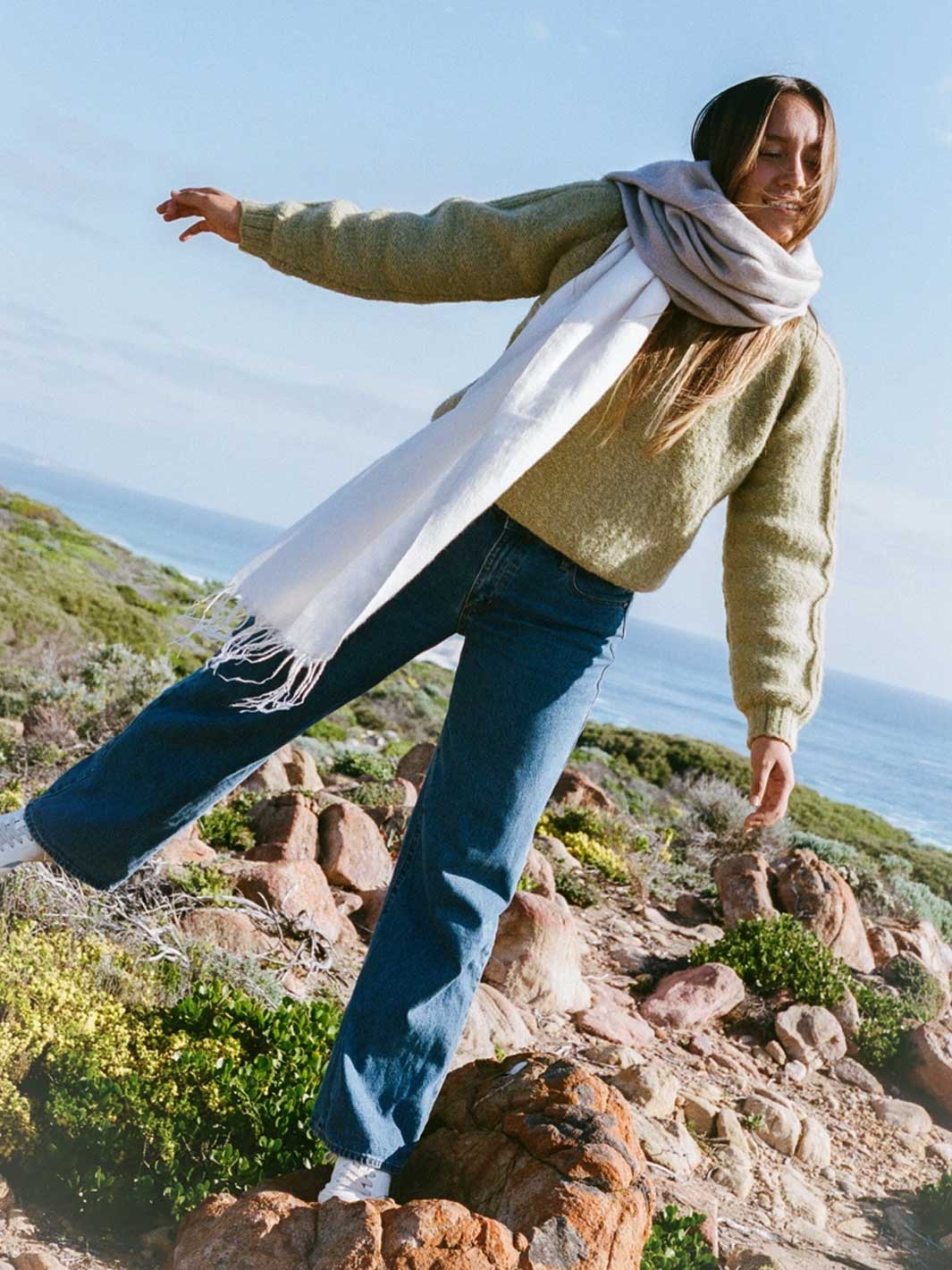 A woman outdoors on a rocky coastal path wearing a textured sage green sweater, blue jeans, white sneakers, and a long taupe and white scarf with fringe, with the ocean and sky in the background.