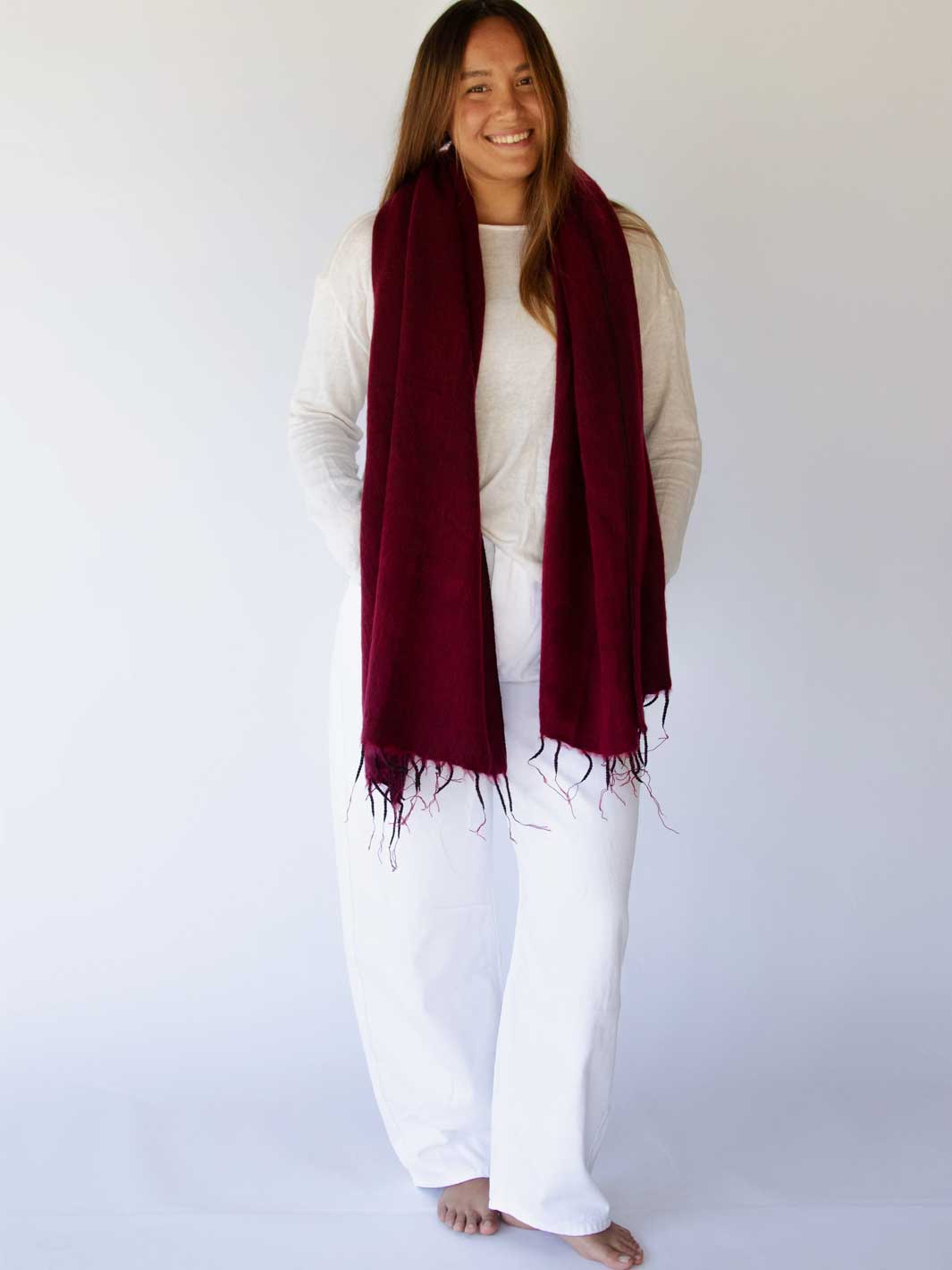 A woman modeling a long, deep burgundy scarf with fringed edges over a white top and white pants, standing against a plain white studio background.