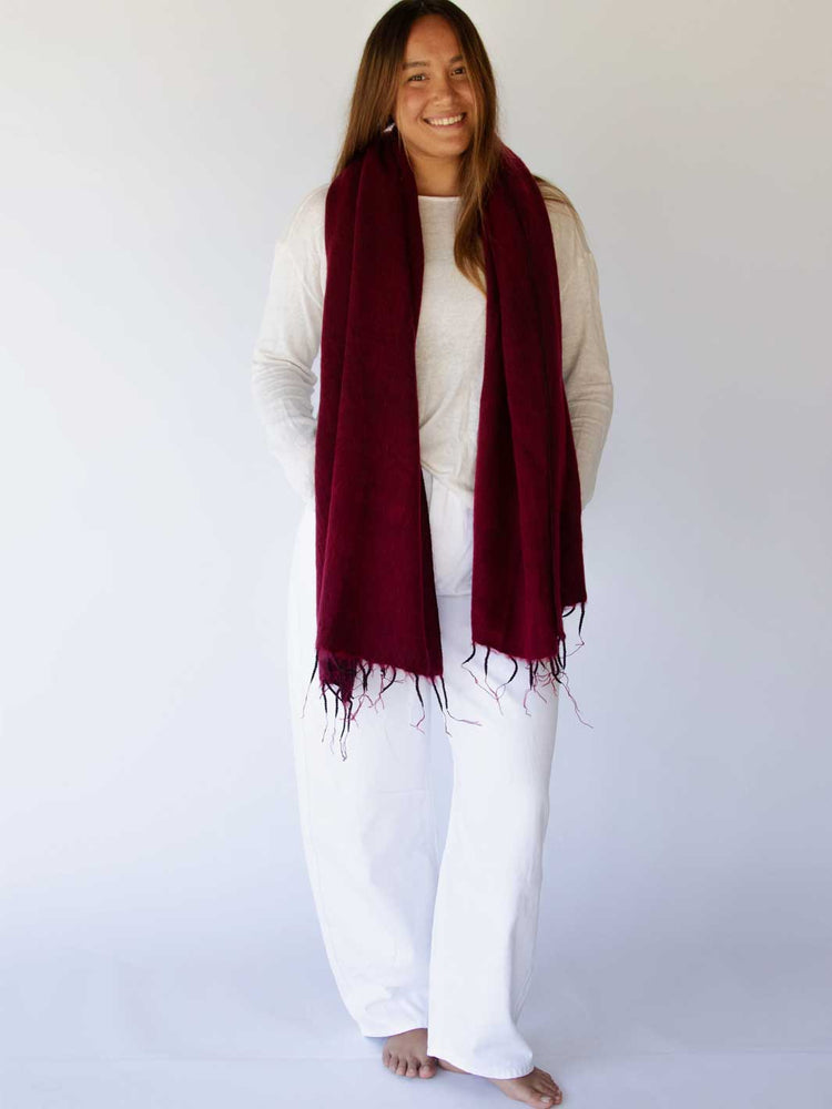 A woman modeling a long, deep burgundy scarf with fringed edges over a white top and white pants, standing against a plain white studio background.