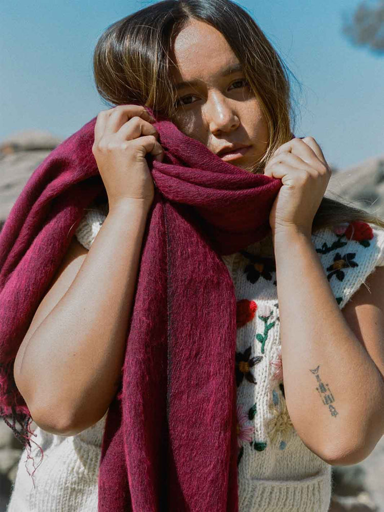 A woman outdoors wearing a chunky knit cream vest with colorful floral embroidery and holding a deep burgundy scarf around her neck.