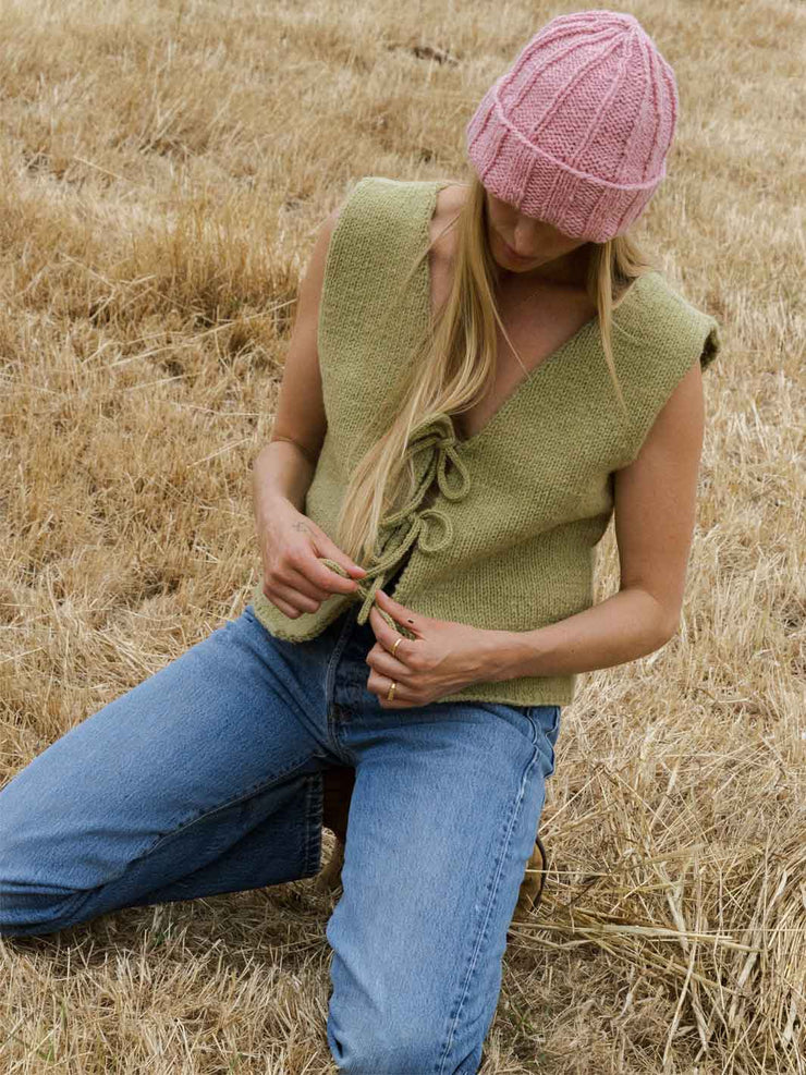 Woman wearing a green knitted vest and pink beanie in a field of dry grass.