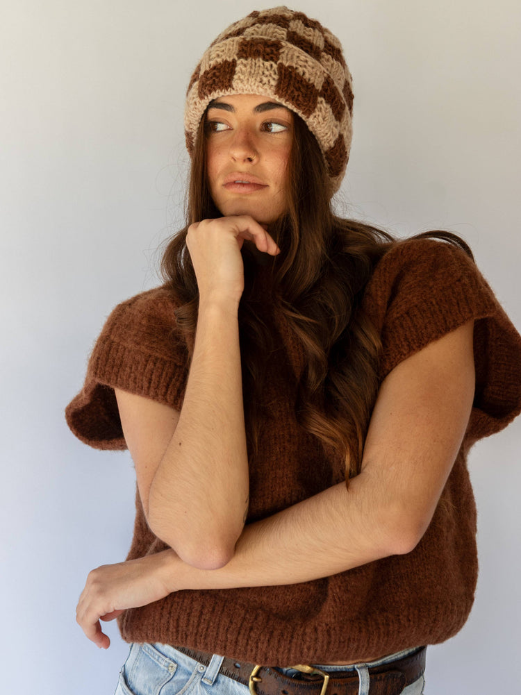 Woman wearing a brown knitted vest and checkered beanie against a white background