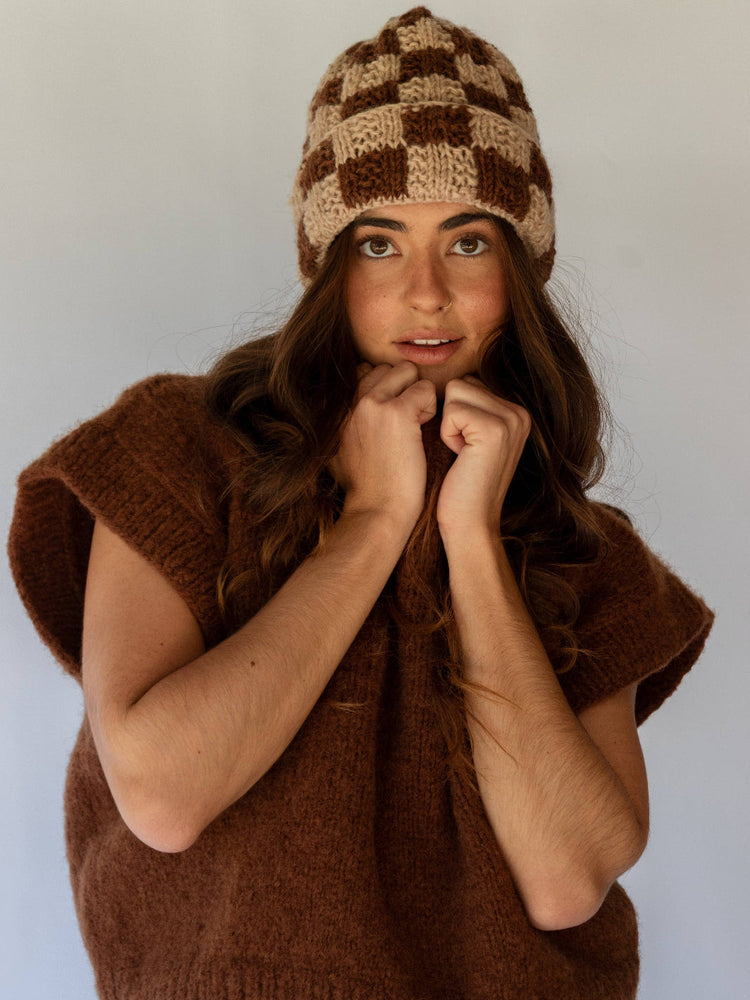 Woman wearing a brown knitted vest and checkered beanie against a plain background