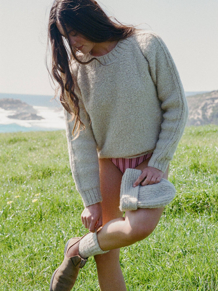 Woman in a beige sweater and shorts standing in a grassy field with a scenic background.