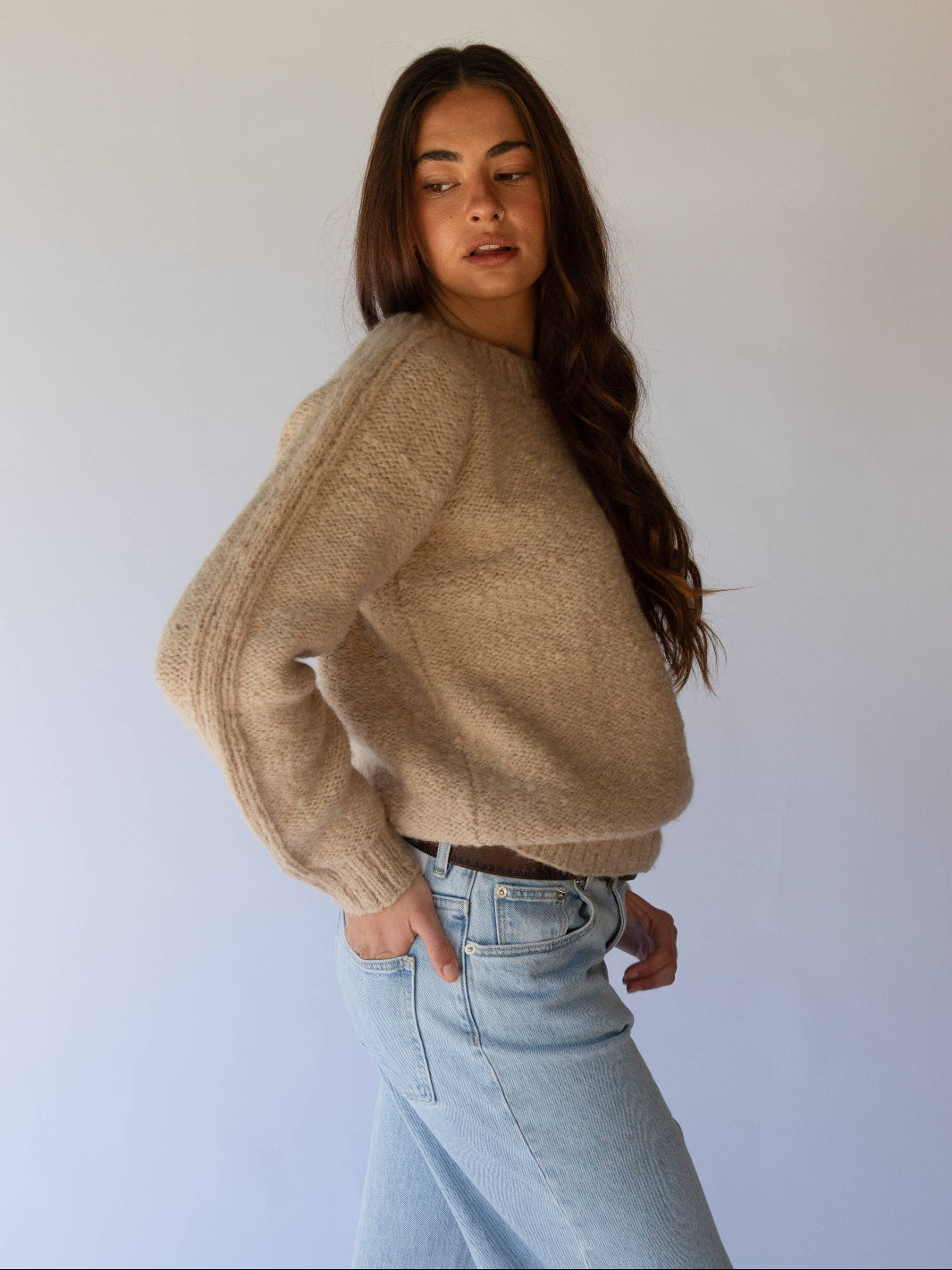 Woman wearing a beige sweater and light blue jeans against a white background