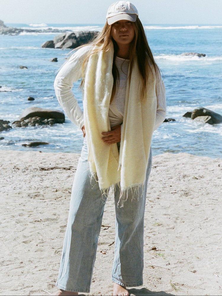 Woman standing on a beach wearing a white cardigan, light blue jeans, and a cream scarf.