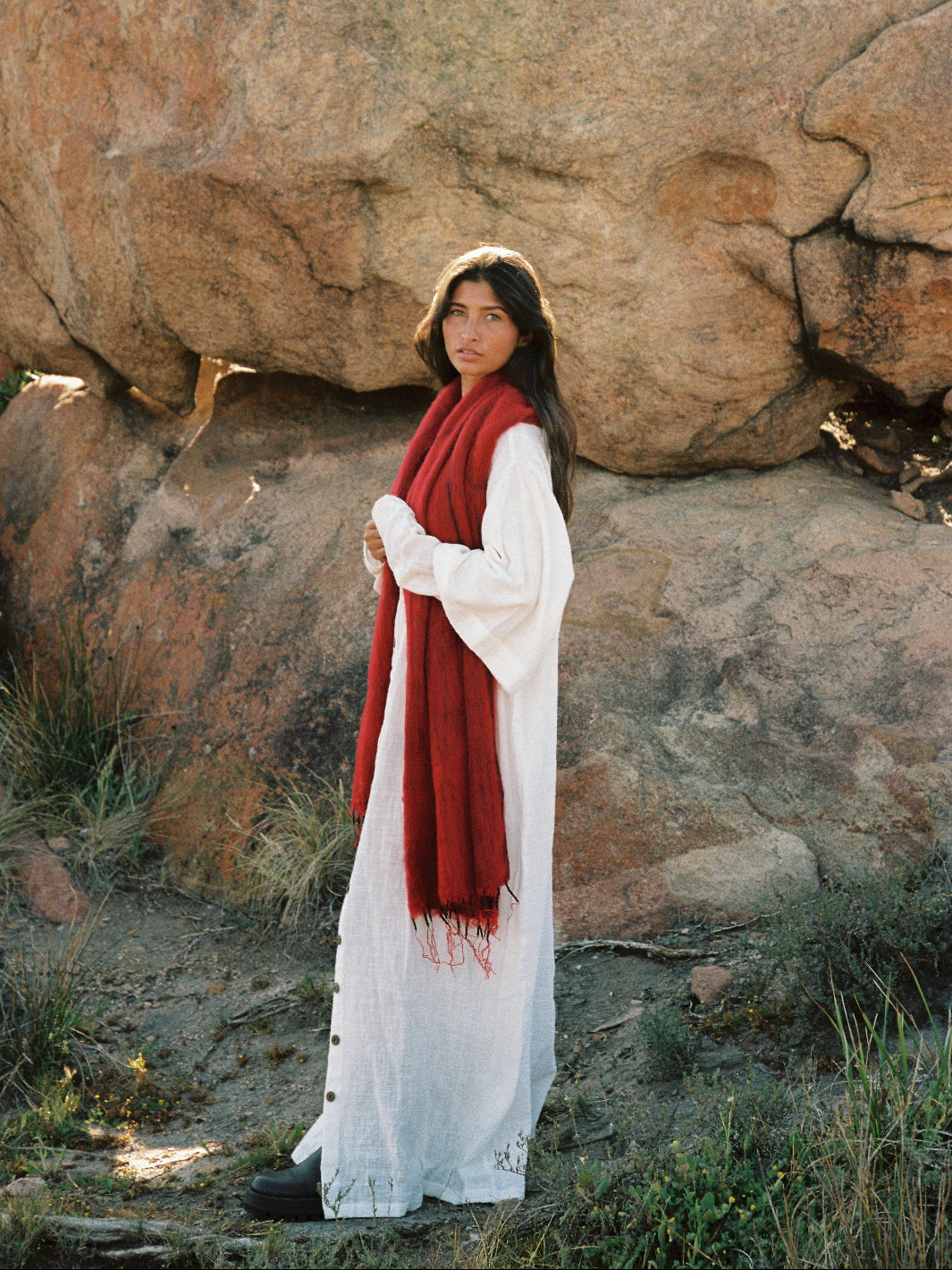 Woman in a white dress with a red scarf standing in front of large rocks.