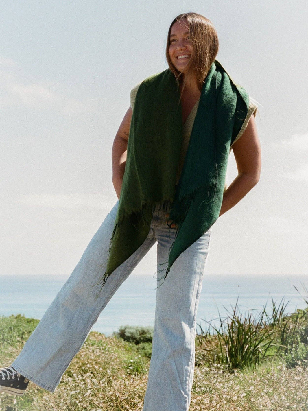 Woman wearing a green scarf and light-colored pants standing outdoors with a clear sky.