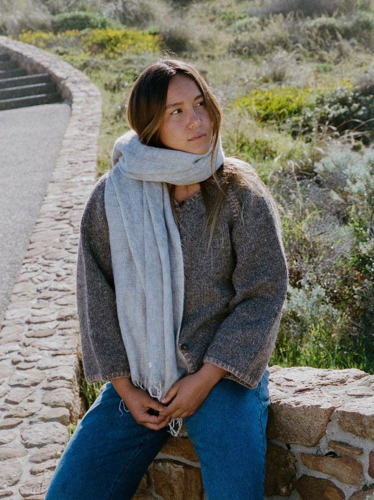 Woman sitting on a stone wall with a scarf and sweater, surrounded by natural scenery.