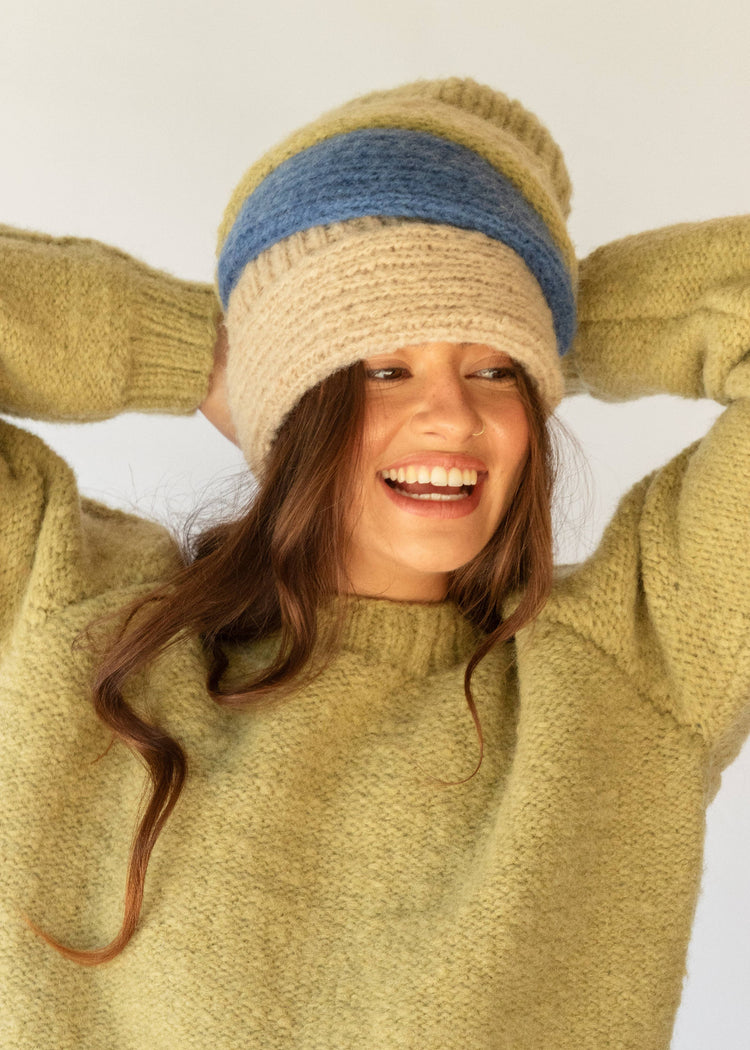 Woman wearing a beige knit hat with a blue band and a green sweater, smiling against a white background