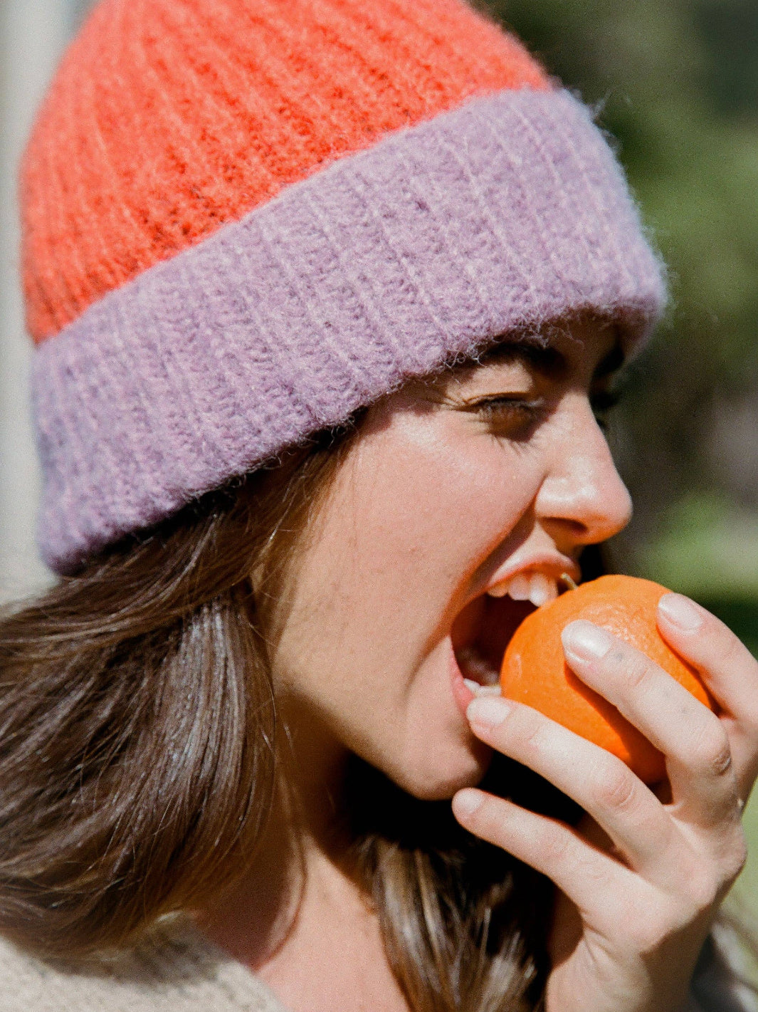 Person wearing a colorful knit hat eating an orange outdoors